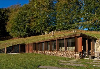 The residents store wood in the facade’s recesses, which, when filled, help the building disappear into the surroundings. The architects used stone sourced from the site for the retaining wall.