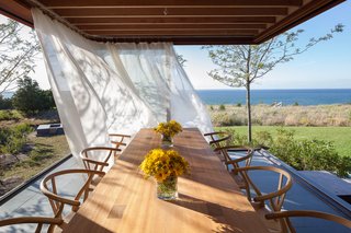 The dining area sits adjacent to the kitchen, defined by three glass walls that open the room completely to the elements, while the roof still provides shelter and shade. A long, family-style dining table made by Larry Hepler anchors the space.