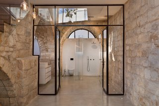 The master bathroom is framed by a glass and varnished steel doorway. Minimal white tiling and concrete floors allow both the original stone walls and graphic geometric glass openings to take center stage.