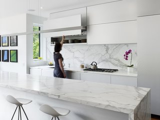 The kitchen contains Onda barstools by Stua from Design Within Reach that surround a 13-foot Calacatta marble island. Custom white aluminum cabinets float above the kitchen appliances by Miele with a stovetop from Wolf and a range hood by Airmec Bello.