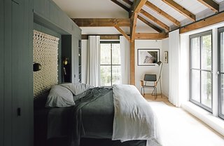 A Passive House and "Sauna Tower" Join a 19th-Century Barn in the Hudson Valley - Photo 4 of 13 - The bedding is intentionally simple, without layers of unnecessary pillows, and the "non-headboard headboard," as Santos describes it, is a loose macramé wall hanging by Sally England. The built-in storage wall that defines the space is painted Kendall Charcoal by Benjamin Moore in a matte finish—a shade used on millwork throughout the house. The Isaac brass sconces are from Schoolhouse Electric, and the floor lamp is from Crate & Barrel. The rocker was custom made by Onefortythree in Las Vegas.