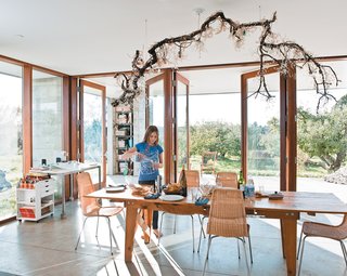 The dining room table sits at one end of the main room, with an open view onto the rows of trees that extend out from two sides of the house. Natasha sets the table underneath a suspended fixture made by her mother, Naomi, out of a salvaged branch, crystal pieces, and strung bulbs.