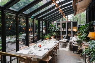 Crawford created a covered patio addition where guests can relax and enjoy a meal within the garden. The leafy glass house holds modern wood furnishings and cozy textiles.