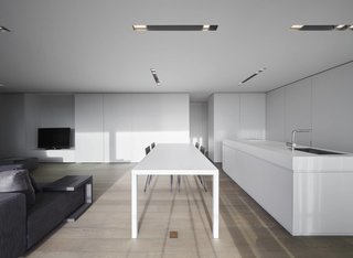 Oostduinkerke is a clean white interior located in West Flanders, Belgium, designed by minus. The space is characterized by a series of strips recessed within the ceiling that conceal lighting. The main living area is open floor plan with the kitchen, dining, and living room combined into one space. The dining table is situated parallel against a large kitchen island with a full service kitchen hidden behind the cabinetry. Sliding doors provide privacy and separation between the various programs.