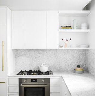 The white minimalist kitchen is fully outfitted with premium Bertazzoni Italia and Bosch appliances. It also features Italian Cararra marble countertops, an island, custom-designed cabinetry, and natural brass Fantini fixtures. 