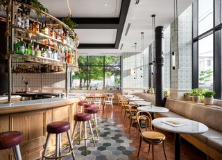 The cafe is designed with plush leather benches and marble tables around its perimeter, as well as black pendant lights hanging above. The flooring marks the separation between the cafe seating and the center bar. 