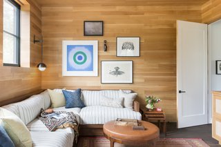 The media room is clad in floor-to-ceiling oak panels. The custom-made, built-in oak banquette was designed by ABD Studio and upholstered in a striped linen. The eclectic mix of artwork is from the collection of the owner and includes pieces from Kiki Smith, Kenneth Noland, and a sculptural work from Mary Shaffer. The rug and side tables are vintage. 