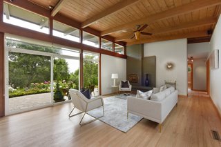 The open-plan living room maintains a strong midcentury vibe and features floor-to-ceiling glazing, tongue-and-groove beamed ceilings, an original brick fireplace, and refinished hardwood floors. 