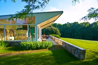A dramatic triangular wooden truss extends the butterfly roof beyond the glass wall of the living room, also shading the stone-paved terrace. A low stone wall expands from the house into the surrounding landscape.