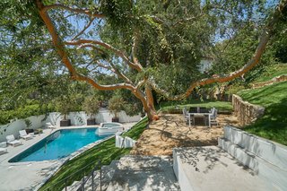 A stone walkway weaves through a grassy, terraced backyard, which is studded with mature trees and complete with a flat lawn, al fresco dining area, and a walled courtyard surrounding an oversized pool, spa, and hills.