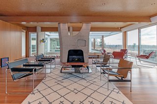 The living room features full-height, wraparound walls of glass and elegant wood details—however, the highlight is a lovely bush-hammered concrete fireplace.