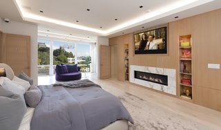 The master bedroom is equipped with two TVs, a marble-framed fireplace, a refrigerator, a microwave, and dual bathrooms—each with their own walk-in closet. Floor-to-ceiling glass doors lead to a private terrace with views of downtown Los Angeles.