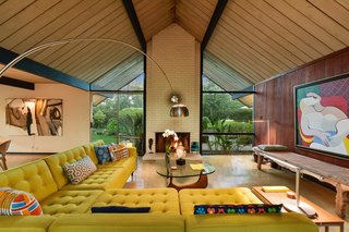 The open-plan living room features classic midcentury elements such as a tall vaulted ceiling, full-height glazing, and a strong brick fireplace.