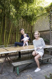 Lara, Ethan, and their baby daughter relax on their back patio, shaded by bamboo.