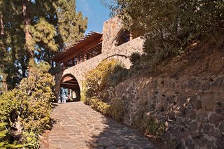 Find Seclusion in the City With This $2.5M Lloyd Wright Residence - Photo 1 of 7 - A driveway passes under an arch and leads to the rear motor court. The exterior facade of the house is made of stone and concrete, with long bands of windows featuring fir mullions and redwood sills. It also had exterior sheetrock installed in a shiplap style, which Powell replaced with marine-grade fir plywood: "The white color was jarring and exaggerated any inharmonious detail."