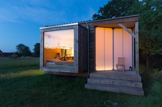 Suspended over the meadow, the bedroom boasts a large glass pane, while the transparent screen door allows the surrounding trees to cast playful shadows across it.