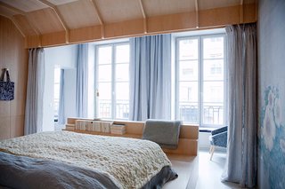 The bed is encased in a birchwood cocoon that looks out to grand French doors that open up to the city beyond. One wall is covered with a floral textile that brings out the calming shades of blue and gray in the space, and the bed area can be partitioned off with floor-to-ceiling curtains.