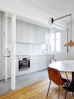 The white walls, ceilings, and decorative crown moldings continue into the kitchen area, where light gray cabinets and a medium gray floor covering highlight the marble of the tabletop and backsplash. 