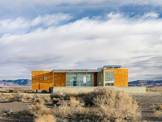 Set in a Las Vegas desert, this prefabricated prototype designed by architect Peter Strzebniok was constructed in sections which were transported, then assembled on site. The house is composed of two complete units with interlocking sections that are raised above the ground to create a clean-lined geometric wood paneled structure of a deep gold color. Within, a "T" shaped floor plan encompasses three bedrooms, and a large living, dining, and kitchen area that connects to a spacious deck. 