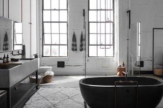 An oversized, custom-made concrete trough on a bronze base is paired with industrial copper piping and taps. The mirror, which is suspended from the ceiling by braided leather straps, was manufactured by PSS Design Cult. The oval bathtub is from NativeStone.