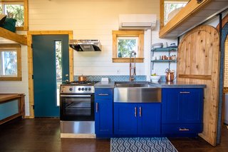 Between the dining area and bathroom is a kitchen that has concrete countertops, a stainless-steel farm sink, an oven and stove, and bright blue cabinets with elegant brass handles. These contemporary touches complement the craftsmanship and industrial-style details, giving this small dwelling a unique, modern feel.