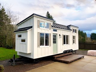 The tiny home rests upon a 28’ x 8.5’ Iron Eagle Tiny House Trailer with three 7K axles, trailer brakes, and road lighting. It has board and batten siding with Pacific Cedar accents, a standing seam metal roof, and a glass exterior door.