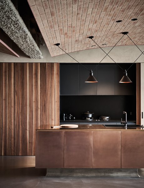 In the open kitchen, wood-paneled sliding doors conceal dark cabinetry, and together with a sleek, dark counter, and a shiny bronze-clad island with a sink, the kitchen becomes part of the overall design rather than simply an area of utility.