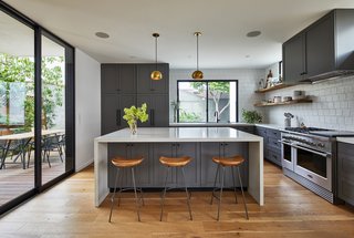 The kitchen is kept light and bright by multiple windows and a sliding glass door, as well as white countertops and a white tile backsplash. The gray cabinets are painted in Benjamin Moore "Kendall Charcoal" paint in semi-gloss finish; the cabinet pulls are "Bowman" hardware by Rejuvenation in Oil Rubbed Bronze finish; and the wood shelves are solid live-edge oak with concealed brackets. 