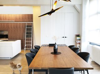 In a condo renovation by architect Samantha Josaphat of Studio 397 Architecture, a kitchen with a generous island opens onto a dining area. The kitchen takes full advantage of the apartment's high ceilings with upper cabinets accessible via a ladder, while the dining area has a sculptural pendant fixture that creates a sense of space and ambiance.