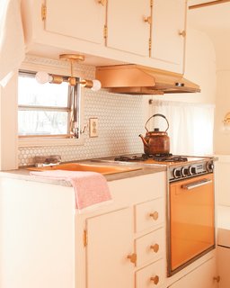 In the petite kitchenette, the original orange-tinted enamel of the sink and stove was spruced up with concrete overlay on the counters, a hex tile backsplash, and new brass accents. The original cabinets were refreshed with the same paint color as the interior walls, then adorned with brass hinges and hex-shaped pulls.