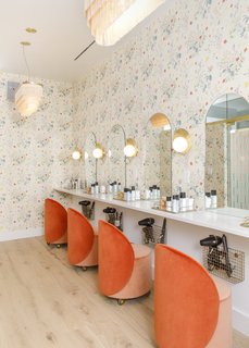In the beauty room, floral wallpaper from Caitlin McGauley picks up the tangerine color of the chairs from Douglas & Bec. Wall light sculptures from Cedar & Moss illuminate the mirrors.