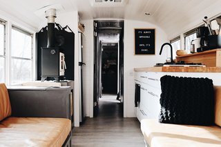 Here is a look at the central aisle to the back of the bus. On the left, there is a fold-up tabletop tucked behind the couch armrest and a seat for one person, serving perfectly as a homework or writing spot. The bus also has a tiny wood stove—along with wood storage—on the ledge to the left of the fridge. 