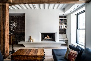 In the living room, the team raised the firebox, cladded the hearth in a tactile plaster finish, and installed a floating limestone bench that wraps the column. On the left (unseen) is integrated firewood storage, and a cozy reading nook sits on the right. "The bench was designed to be used as a social space/lounge, and is well-used," says Coffey. The wood beams and red brick were scraped and stripped many times to remove the silver paint and reclaim a natural state. 