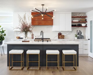By removing walls and a counter peninsula jutting into the room, interior designer Corine Maggio was able to create enough space for a generous island. The stove wall is a fitting focal point with a hood vent accented in tigerwood and a quartz slab backsplash. Black and brass accents, including the Renwil chandelier and Nuevo Living stools, are a theme throughout.