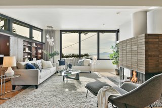Built-in wood shelving sits below clerestory windows opposite a large brick fireplace with a sculptural chute. Expansive windows provide views of the bay beyond. 