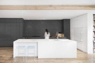 The exterior wall of the kitchen reveals the original brickwork. Gray, full-height, seamless cabinets match the hue of the gray brick. Two light blue counter stools add a pop of color.