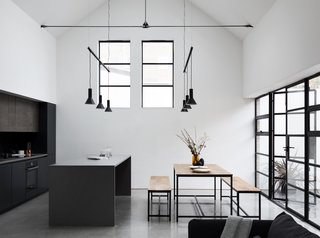 Large steel framed windows and doors frame the open kitchen and dining space. Modern pendant lights hang above each cooking and eating surface.