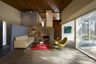 Flagstone flooring flows between the open living spaces. A stone fireplace anchors the main living space, while cedar wood decking extends between spaces above. A blue entry partition adds a pop of modern color.