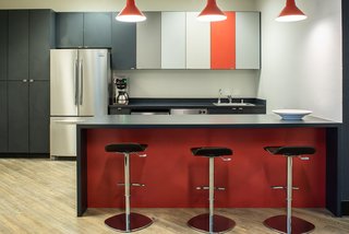 Simple Caesarstone countertops are accented by pops of color in the red bar, multi-tone cabinets, and pendant light fixtures by Troy. Complete with Lez swivel bar stools, the break room is a great place to enjoy lunch or hold an informal meeting.