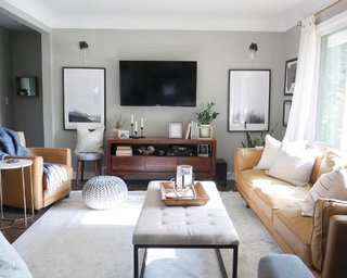 The living room features contemporary furnishings including a Babette brown leather and steel frame ottoman by Christopher Knight Home from Overstock, a Penelope round tufted accent stool from All Modern, a faux marble and gold end table from Pier 1, and a knitted silver-grey pouf from CB2.