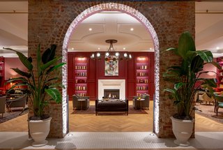 An original brick wall with an arch opening frames views of The Press Room, a cozy lounge lined with paneled walls painted a deep cranberry hue to match the Press Room bar front.