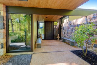 The covered entrance walkway that leads to a oversized bronze door is bordered by a long granite wall on one side and open woodland on the other.