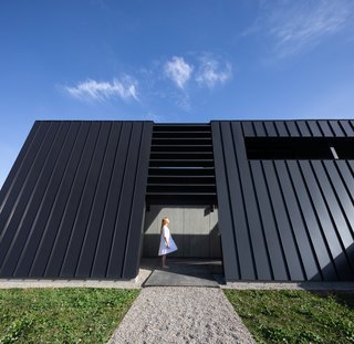 A view of a gravel walkway and the entrance that leads to a covered pathway. "The entry side is hyper minimal and mysterious with slight glimpses out from within, providing only a tease on approach from the road," says the firm.