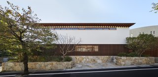 The home is concealed behind horizontal bands of materials including Aji stone quarried from Shikoku, a powder-coated aluminum screen (lath), the white plaster exterior of the second floor, and a titanium zinc alloy roof.
