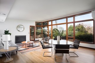 A wall of windows floods the living room and dining area with natural light. The room has been staged with Knoll dining chairs by Mies van der Rohe, a Platner Wire dining table, a Plycraft Lounge chair, and other vintage period-appropriate finds.