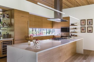 A waterfall-edge quartz-topped island creates a dramatic statement in the minimalist kitchen. A window cutout behind the sink overlooks forest views to the west.