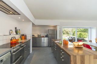 The recently renovated kitchen is now spilt into two sections and features industrial stylings, giving it the look of a commercial kitchen. The deep orange accessories, along with the wooden worktop on the island, brightens the overall gray-toned space.<br>